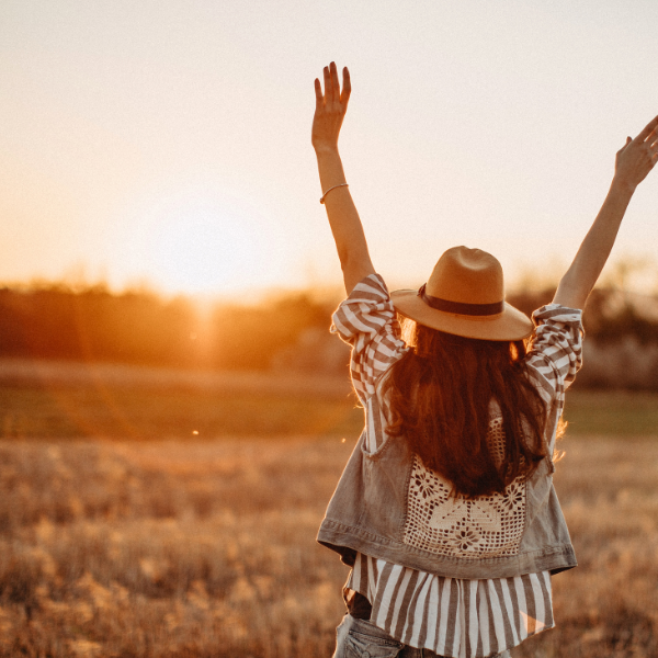 image: A woman standing in a field, facing away from the camera with her arms up in the air. She's wearing a fedora and loose shirt with a vest, giving her a relaxed and triumphant feel. Image represents day one sober, and feeling confident and comfortable.