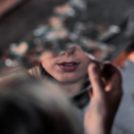 image: Woman holding a piece of broken glass, looking at her reflection. You can only see her nose and lips in the glass. The background is blurred.