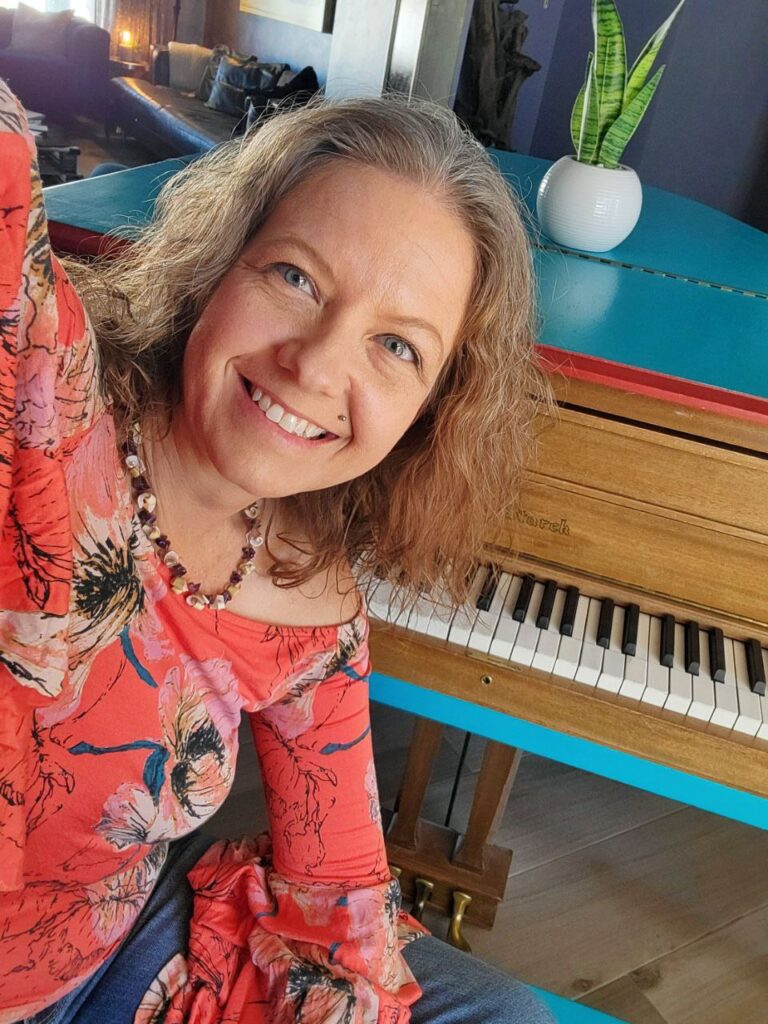 Image: The author, wearing an orange floral shirt and shell necklace, sitting on the stool of a turquoise piano. Image represents the author's battle with self worth and high achieving.